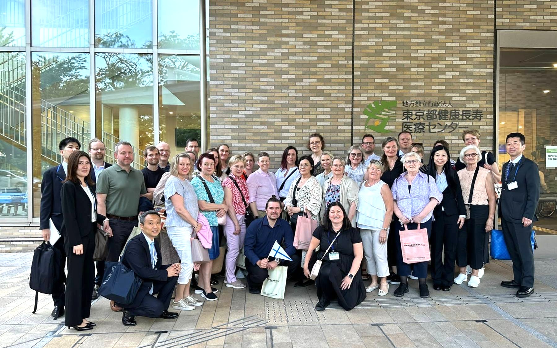A large group of people posing together in front of the Tokyo Metropolitan Institute for Geriatrics and Gerontology building.
